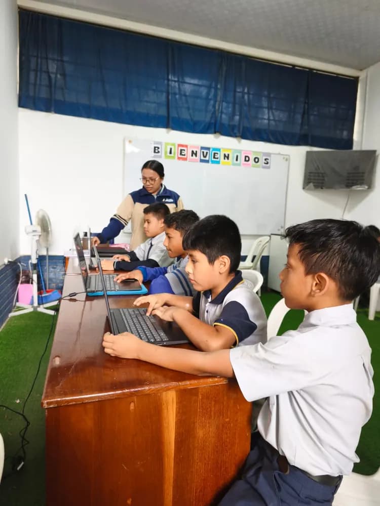 Alumnos trabajando en laptops en el aula de cómputo, acompañados por su docente.